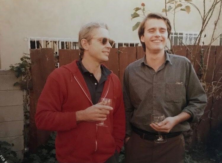 Two men smiling and holding wine glasses outdoors in front of a wooden fence with plants, as featured in RECORD by George Keenen.