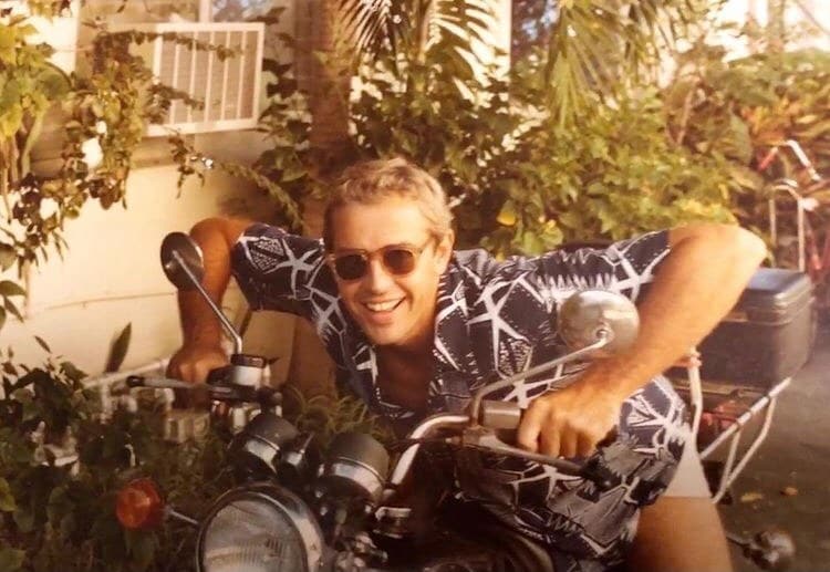 Wearing RECORD by George Keenen, a person smiles while sitting on a motorcycle, surrounded by greenery.