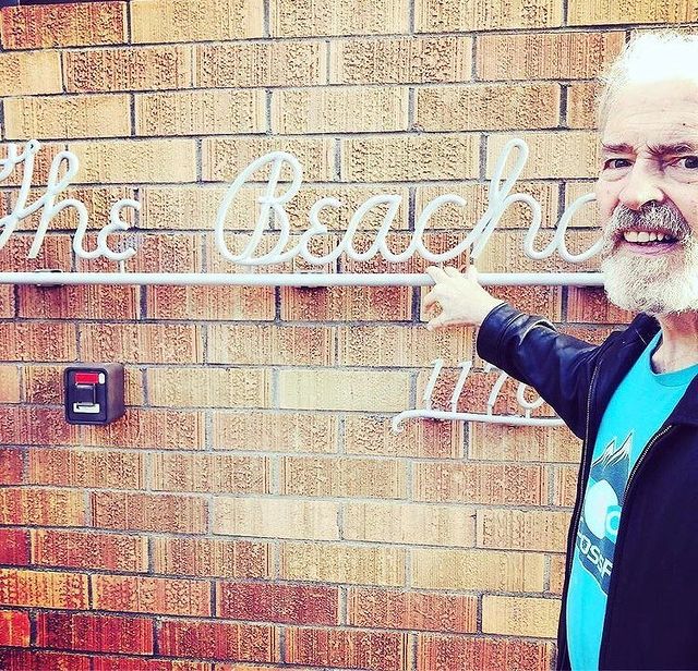 Man with a beard pointing to a neon sign on a brick wall that reads "RECORD, by George Keenen.