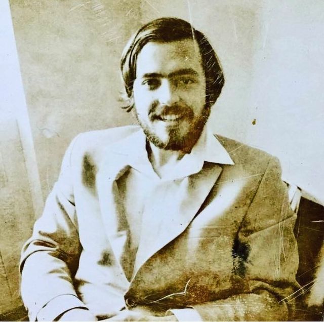 A monochrome image from "RECORD," by George Keenen, featuring a bearded man in a suit, smiling as he sits.