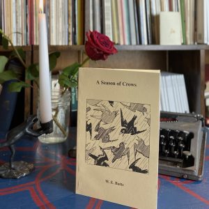 A book titled "A Season of Crows" by W.E. Butts, published in 2000, is displayed in front of a typewriter, candle, and red rose, with a bookshelf in the background.