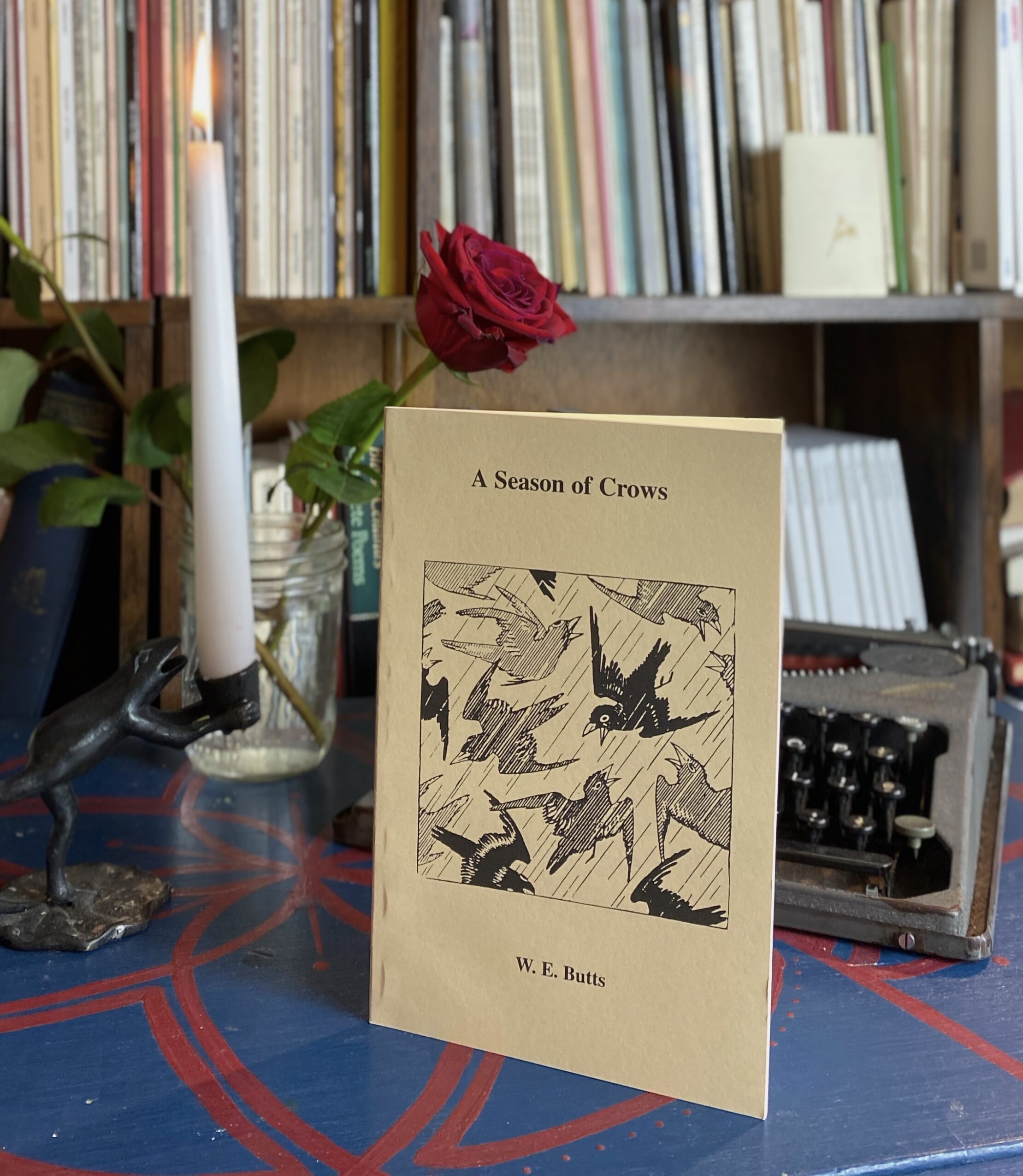 A book titled "A Season of Crows" by W.E. Butts, published in 2000, is displayed in front of a typewriter, candle, and red rose, with a bookshelf in the background.