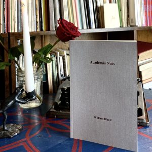 The 2016 book "Academia Nuts" by William Bland rests on a table adorned with a rose in a glass jar, a lit candle, and surrounded by various other books.