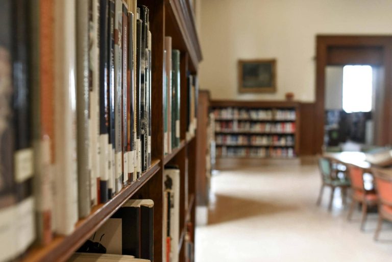 A library aisle with shelves of books on the left and tables with chairs in the background.