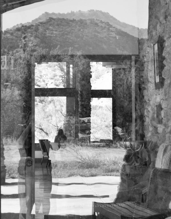 A person photographing through a glass window reflects an outdoor scene with mountains and a stone structure.