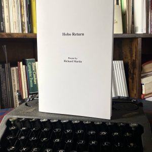 The poetry collection "Hobo Return, Poems by Richard Martin (2021)" is placed on a vintage typewriter, with a backdrop of a bookshelf filled with various books.