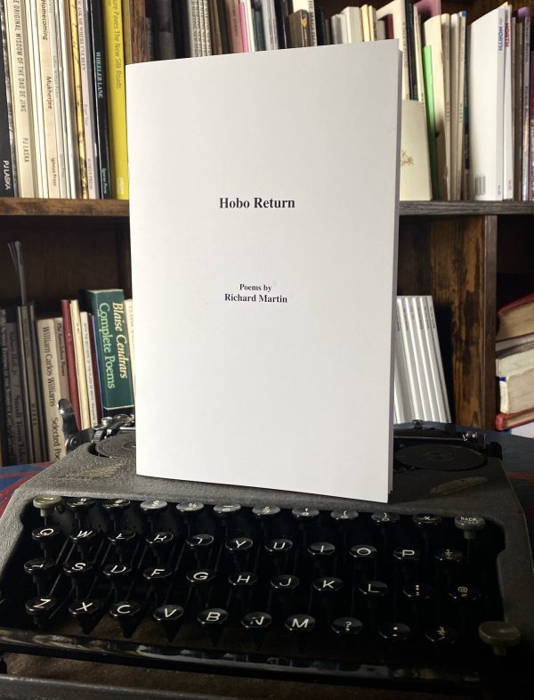 The poetry collection "Hobo Return, Poems by Richard Martin (2021)" is placed on a vintage typewriter, with a backdrop of a bookshelf filled with various books.