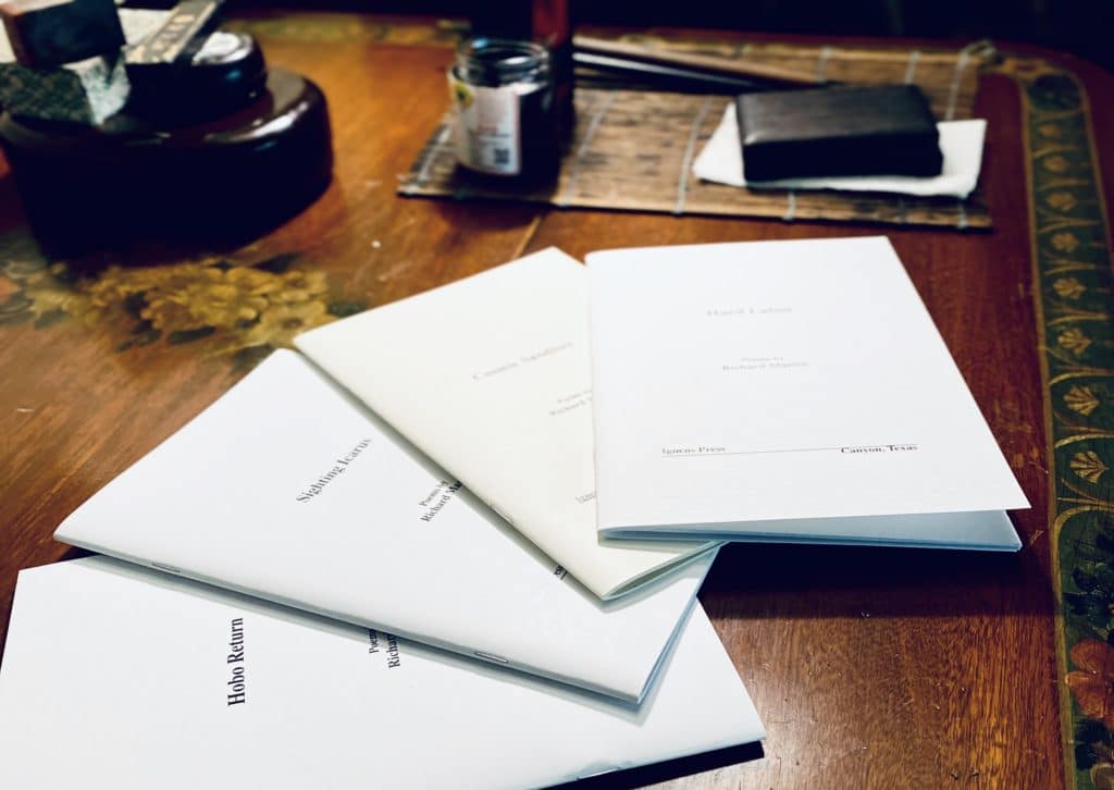 A table with five neatly stacked booklets, each with simple white covers, alongside a notebook and a jar in the background.