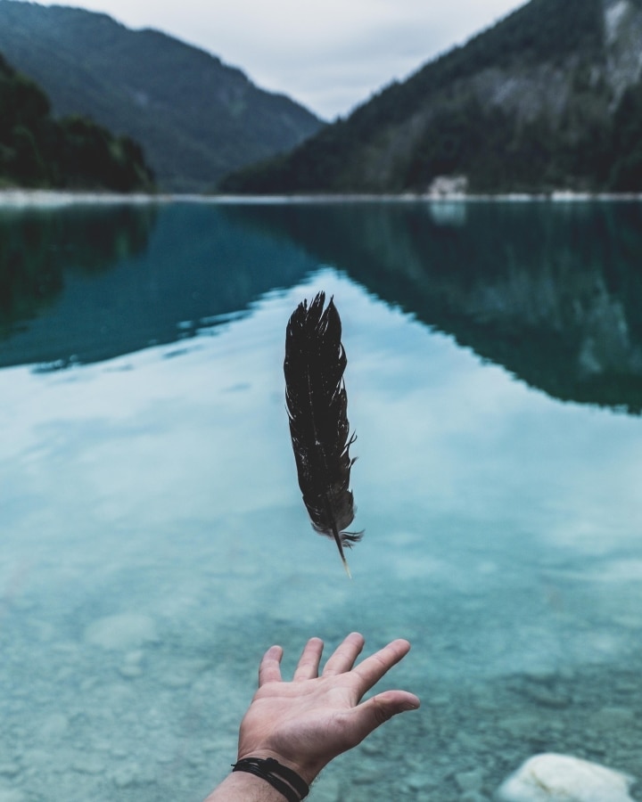 A hand tosses a floating black feather over a clear lake with mountains in the background.