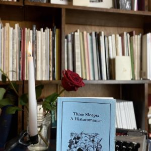 A copy of "Three Sleeps: A Historomance" by Richard Blevins, published in 1992, is placed on a table alongside a red rose, a candle, and a vintage typewriter. A bookshelf brimming with books can be seen in the background.