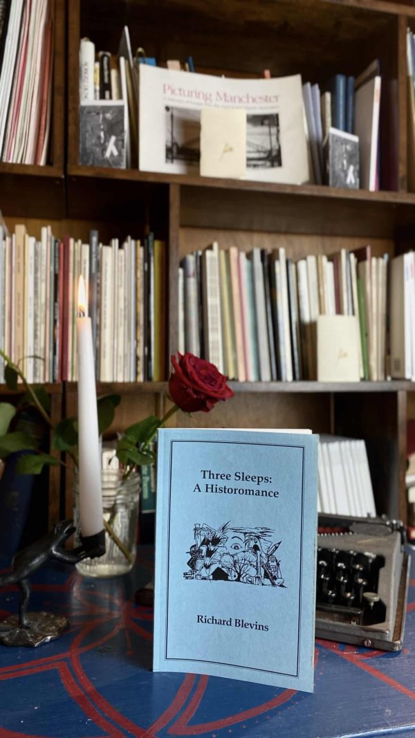 A copy of "Three Sleeps: A Historomance" by Richard Blevins, published in 1992, is placed on a table alongside a red rose, a candle, and a vintage typewriter. A bookshelf brimming with books can be seen in the background.