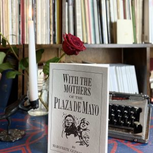 The product, "With the Mothers of the Plaza de Mayo" by Marguerite Guzman Bouvard (1993), is showcased on a table accompanied by a lit candle, a rose, and a typewriter, with shelves filled with books visible in the background.