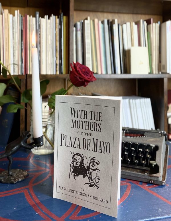 The product, "With the Mothers of the Plaza de Mayo" by Marguerite Guzman Bouvard (1993), is showcased on a table accompanied by a lit candle, a rose, and a typewriter, with shelves filled with books visible in the background.