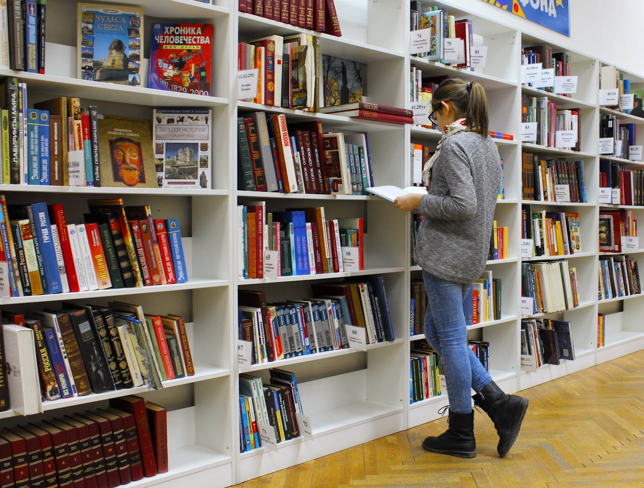 A person stands and reads a book in front of a bookshelf filled with various books in a library setting.