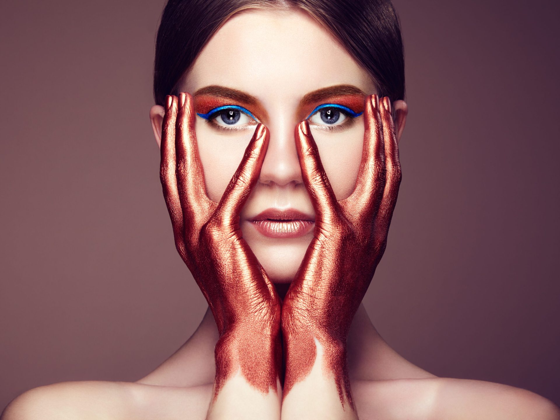 A woman with metallic copper-painted hands framing her face, wearing blue eyeliner and orange eyeshadow, against a beige background.