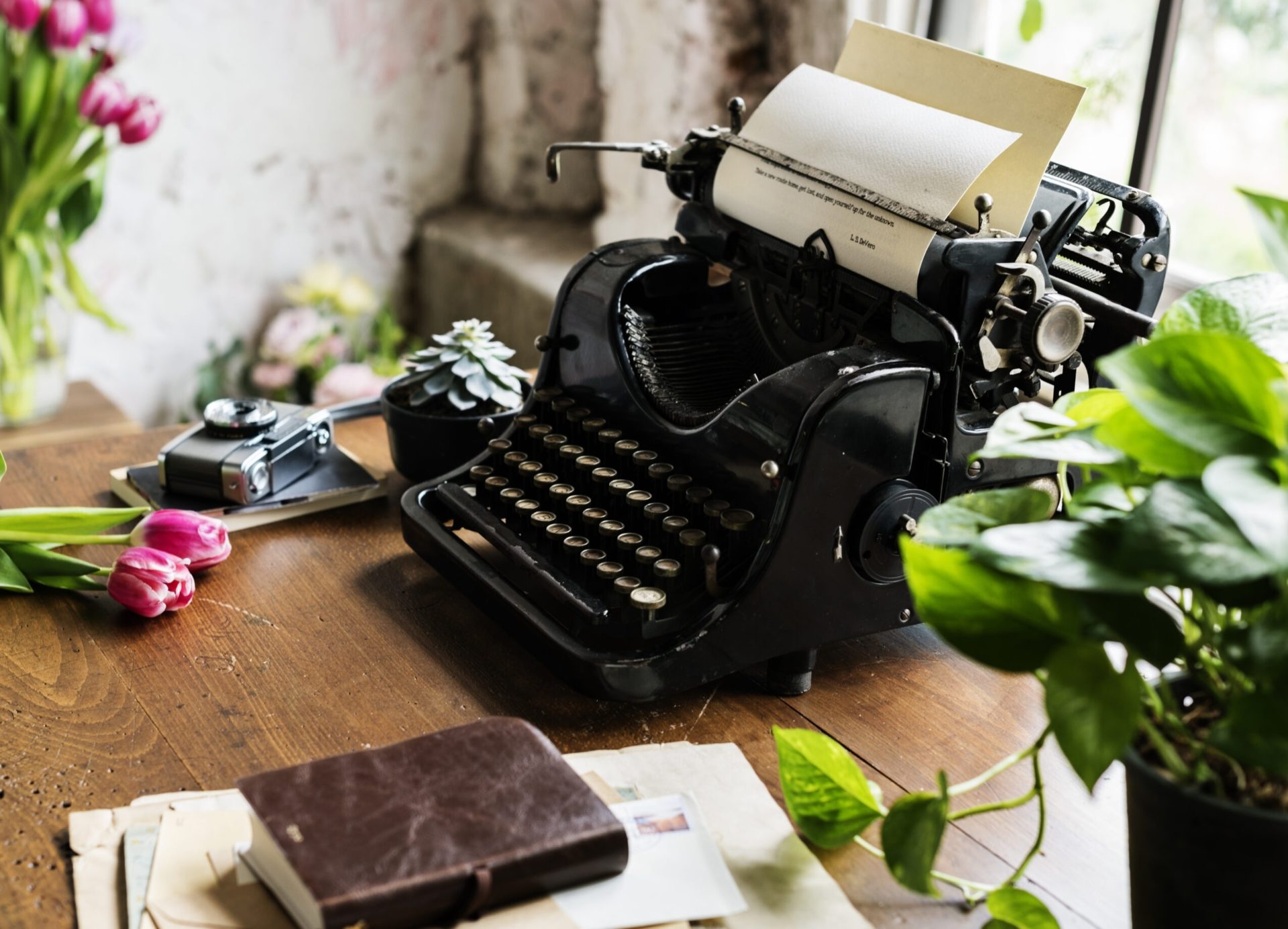 Vintage typewriter on a wooden desk with a camera, notebook, potted plants, and tulips in the sunlight.