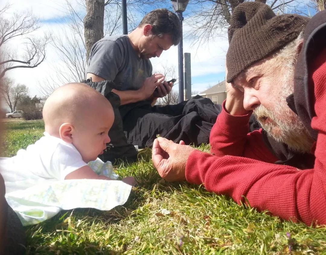 A baby lying on grass looks at an older man holding a small flower, while another man in the background uses a smartphone.